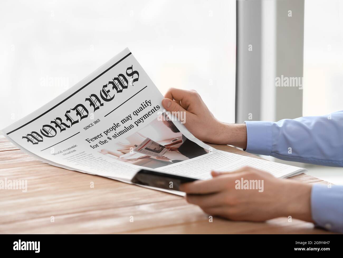 Young man with mobile phone reading newspaper at home, closeup Stock Photo