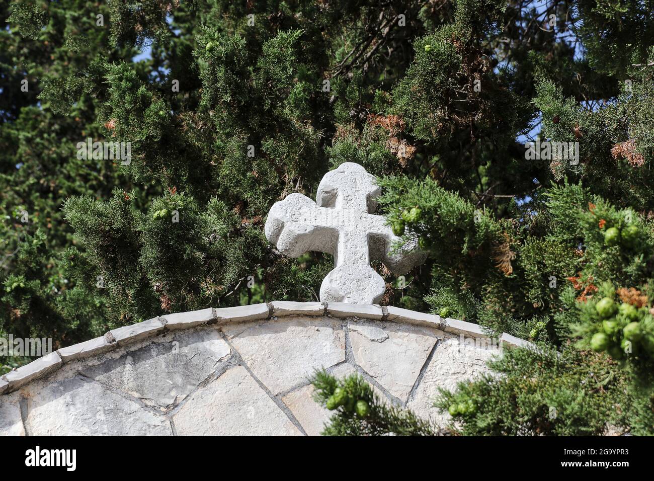 White Christian cross on the top of the stone wall of the church against the green coniferous trees. Stock Photo