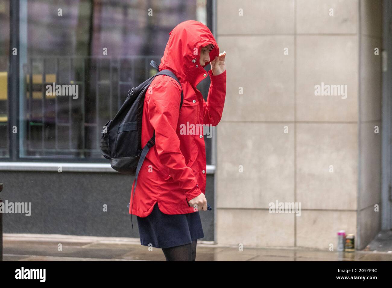 Woman in red anorak holding hood in Preston, Lancashire. UK Weather 28 July 2021; Early morning rain in the city centre with further heavy showers expected.   Credit MediaWorldImages/AlamyLiveNews. Stock Photo