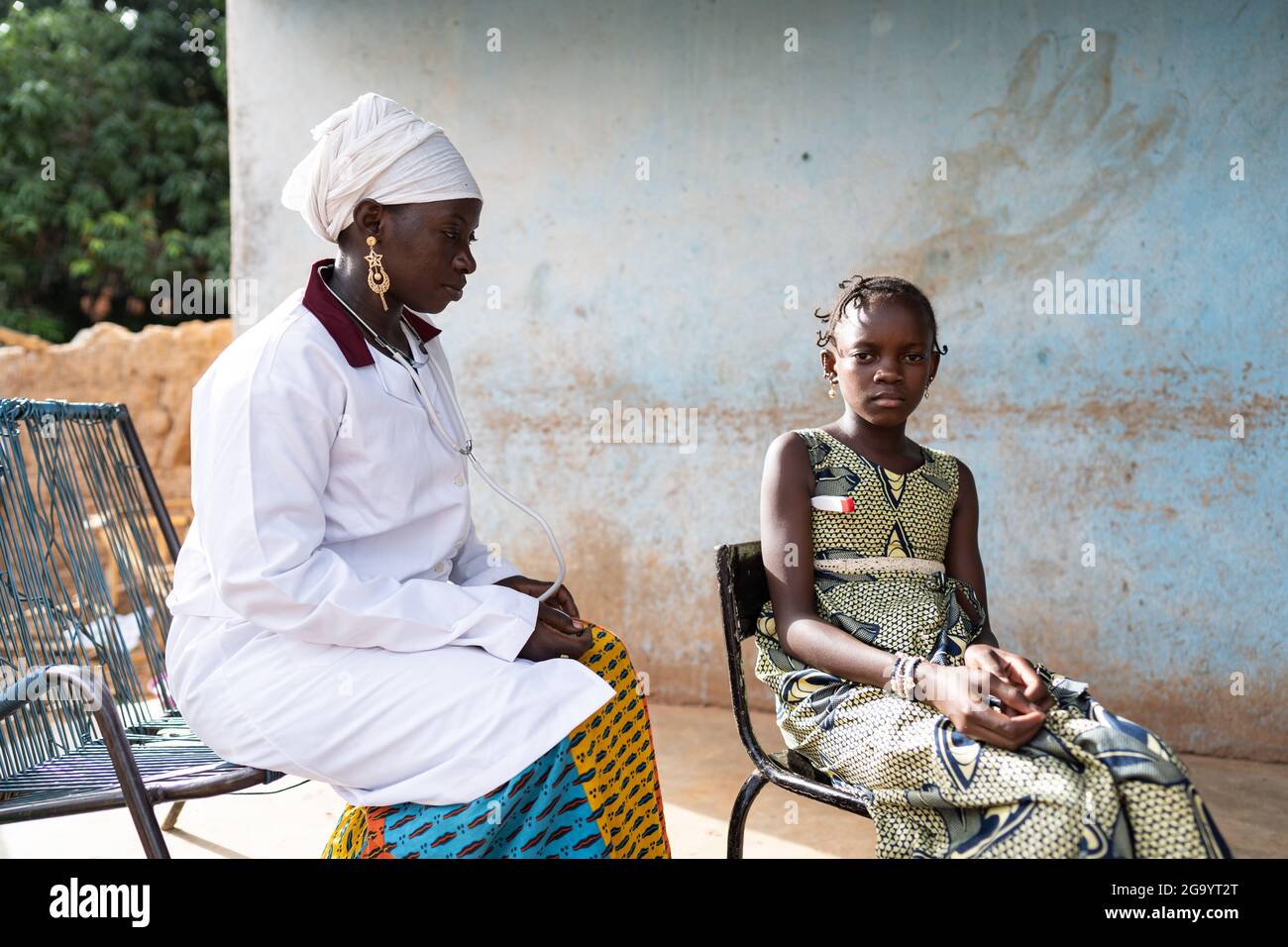 In this image, a sitting black female nurse with hands folded is watching a small African girl sitting at her side with a thermometer under her arm, b Stock Photo