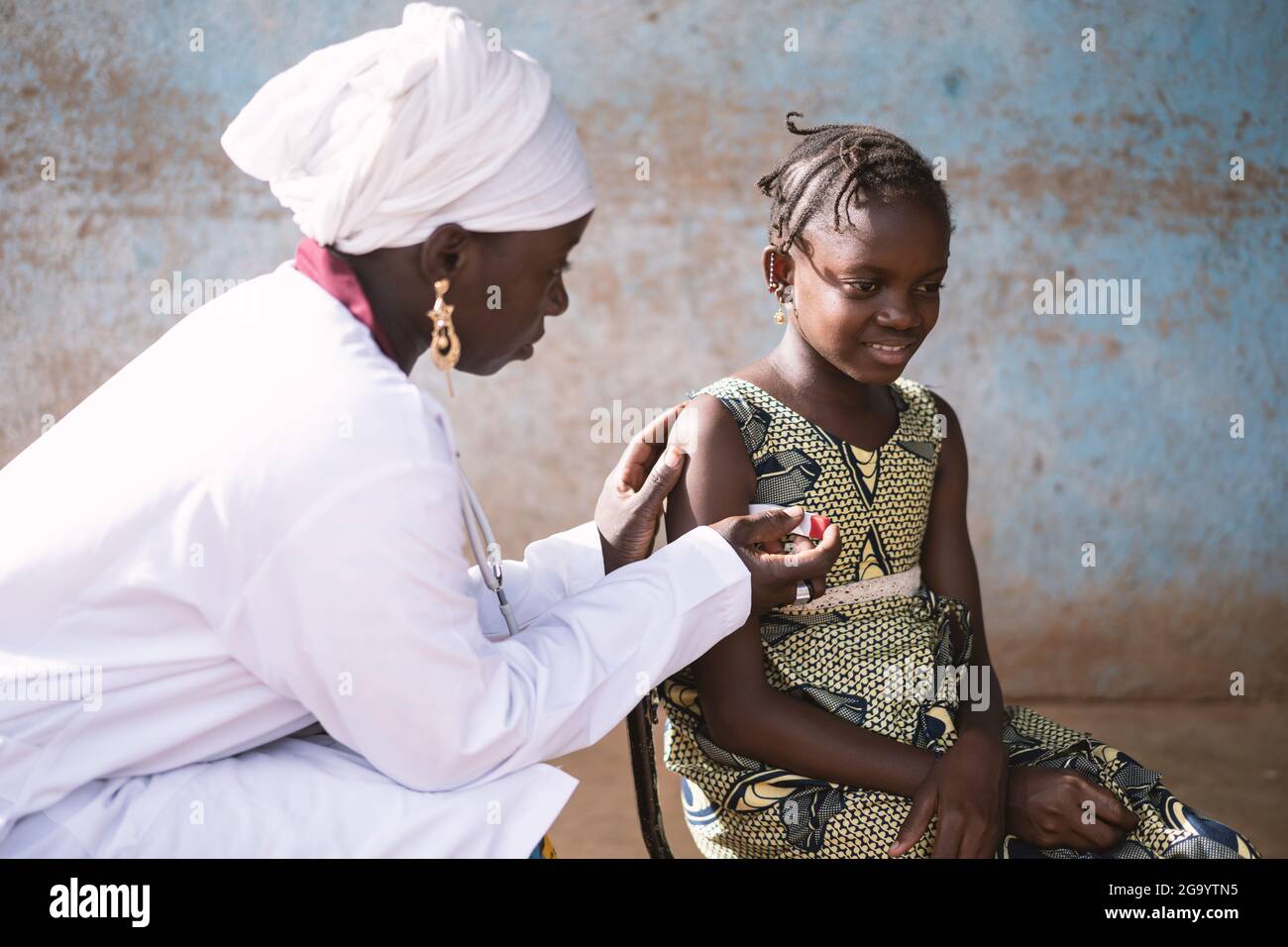 In this image a black female nurse is gently placing a modern digital thermometer under the arm of a smiling little African schoolgirl with fever, in Stock Photo