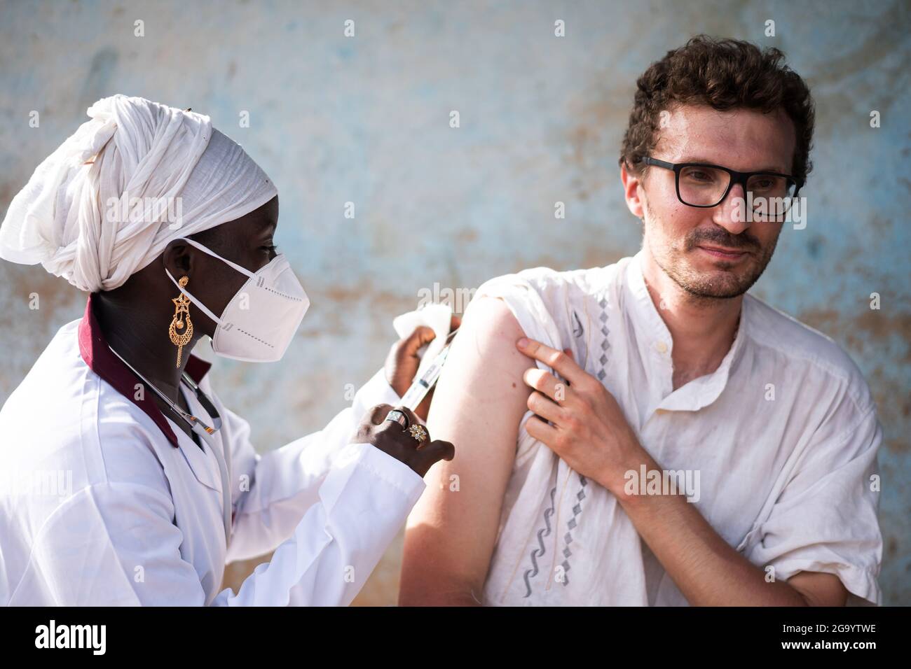 In this image, a young caucasian adult with a tense expression on his face is injected a vaccine shot in his right arm from a black nurse wearing a fa Stock Photo
