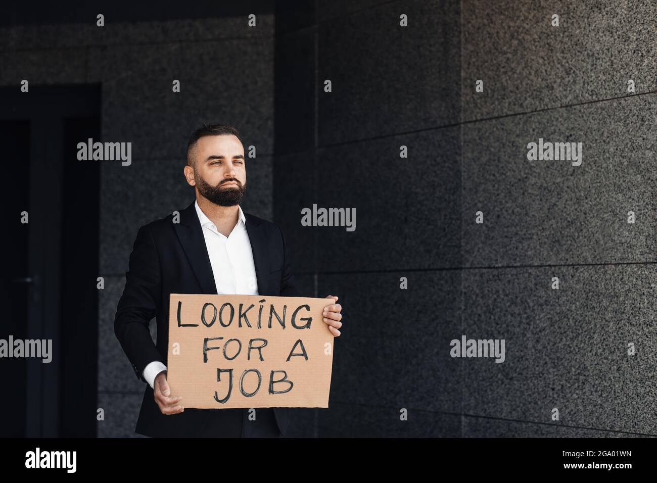 Unhappy despaired businessman lost work holding looking for job placard, standing near business center, free space Stock Photo