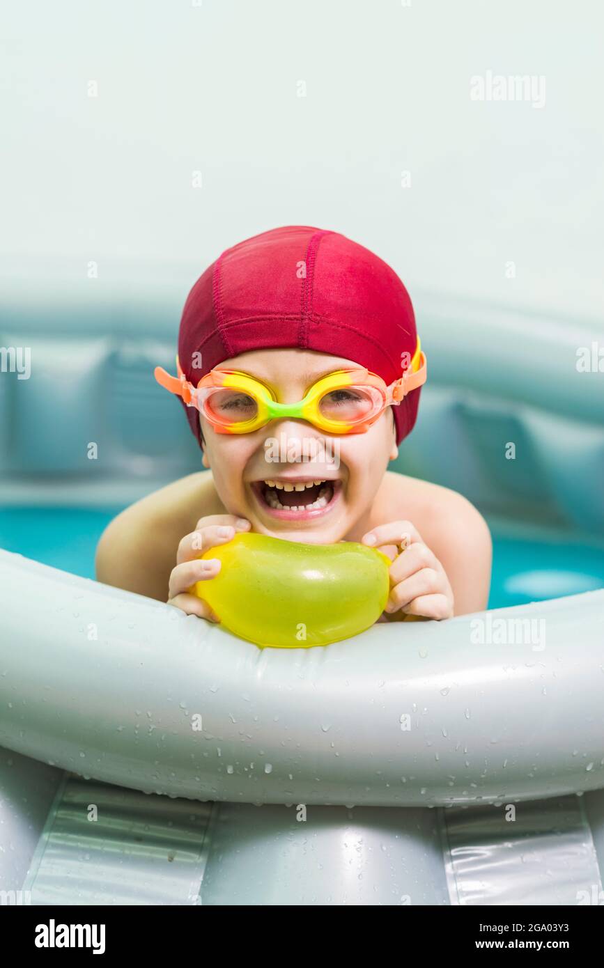 Portrait of a smiling child with a red swimming cap, in a small rubber pool, playing with a yellow water balloon. Summer is the ideal time for water g Stock Photo