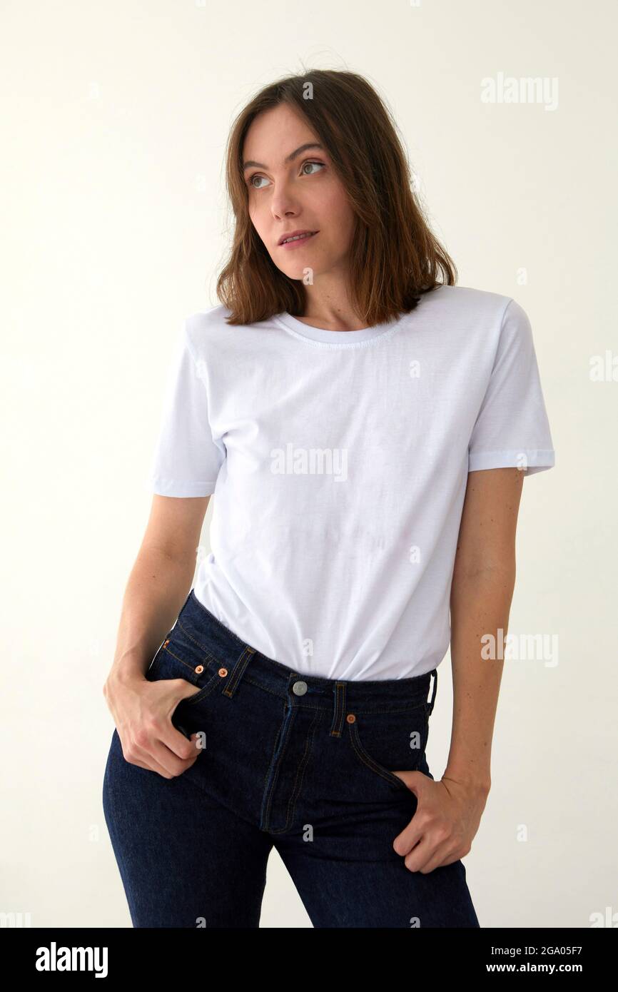 Positive female model wearing white t shirt and jeans standing with hands on waist against white background and looking awey Stock Photo