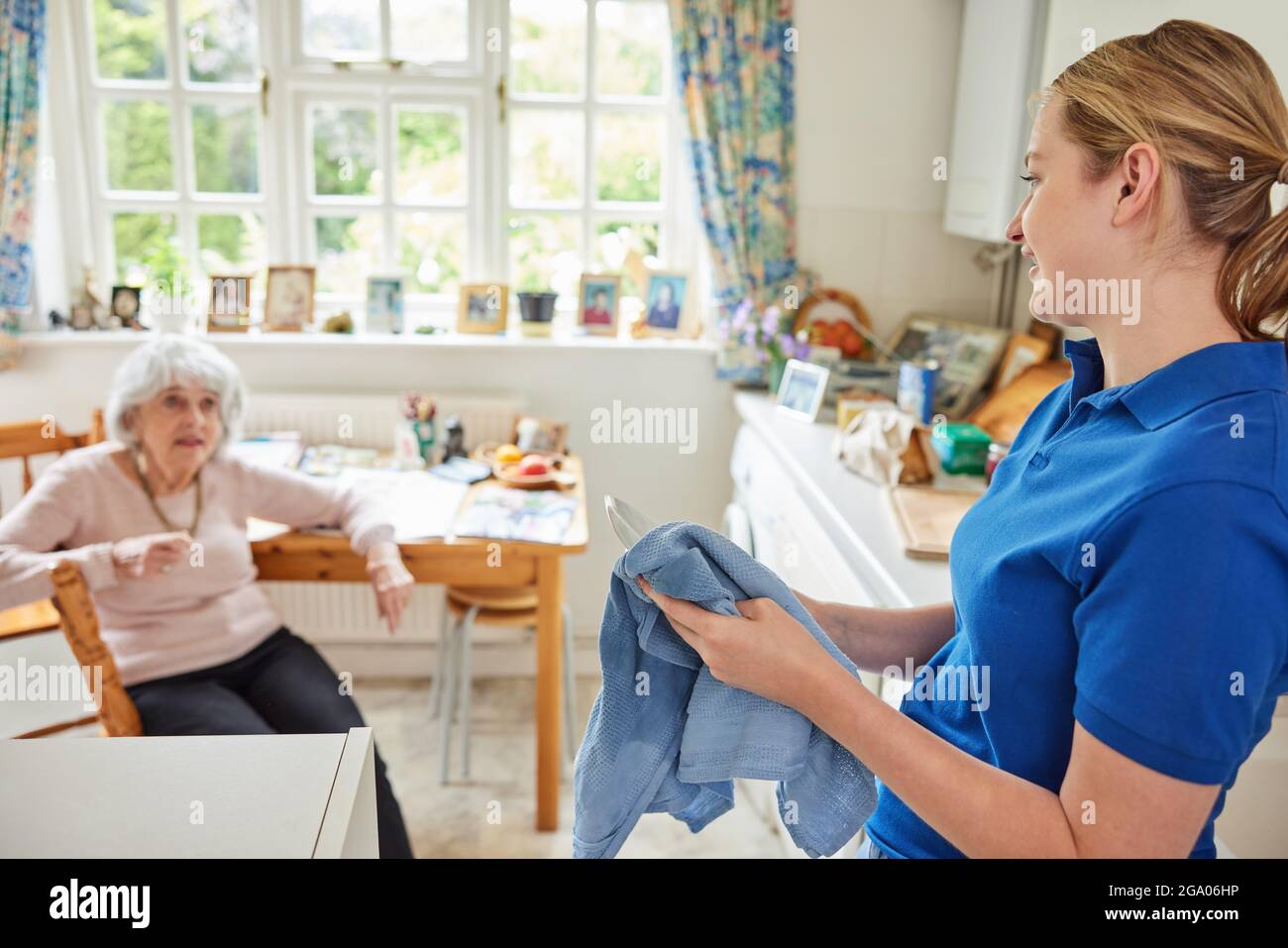 Female Home Help Cleaning House Doing Washing Up In Kitchen Whilst Chatting With Senior Woman Stock Photo