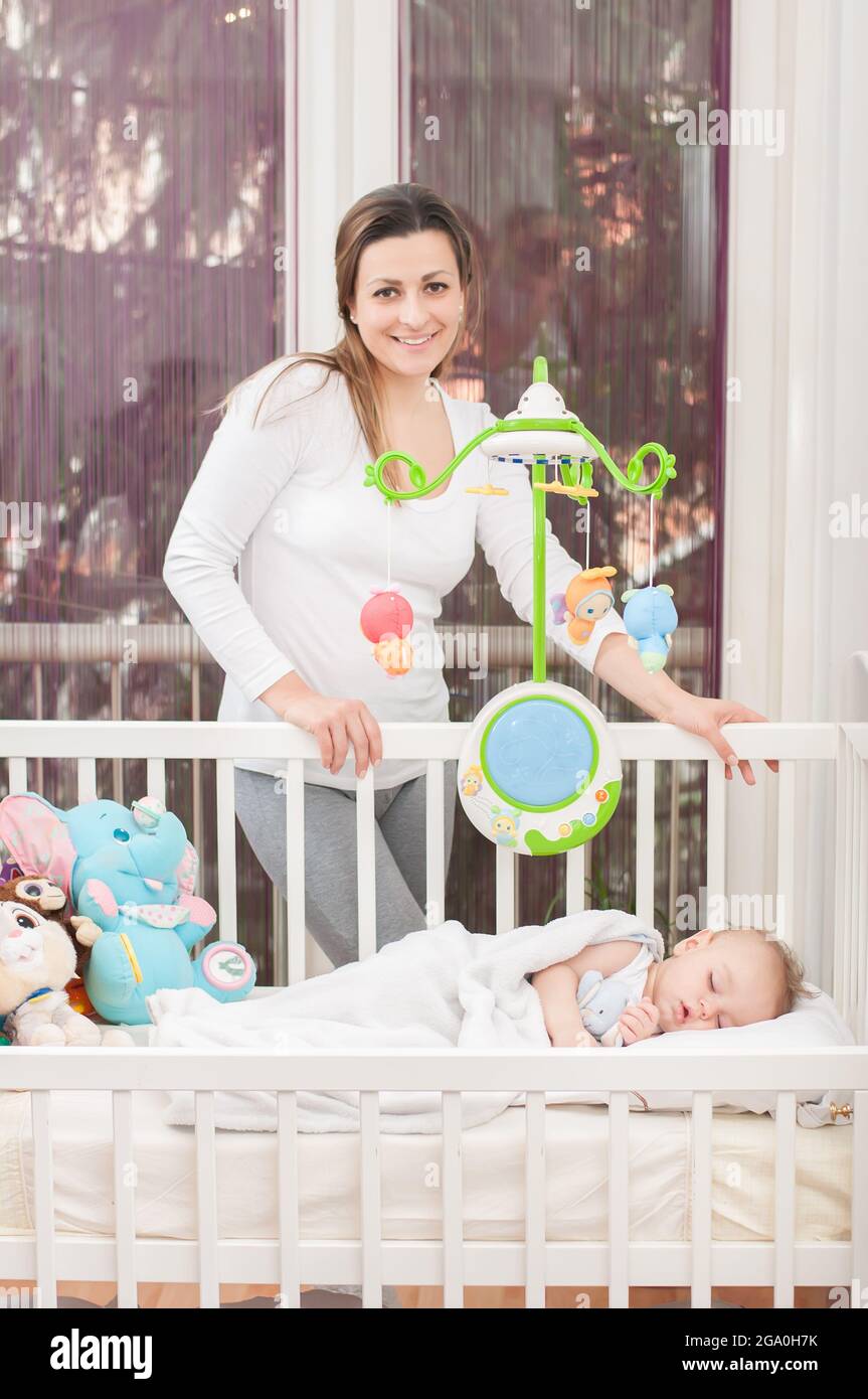Happy mother standing beside the baby's bed and smiling Stock Photo