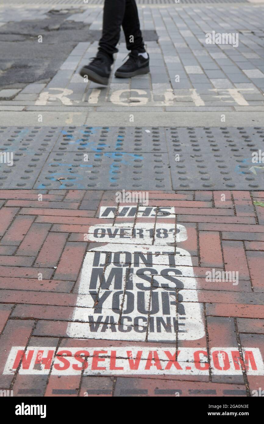 London, UK, 28 July 2021: In Windrush Square, Brixton, a painted sign on the pavement at a pedestrian crossing reminds anyone over the age of 18 that they are eligible for a coronavirus vaccination. Vaccine take-up is lower in big cities than in the rest of the country. Anna Watson/Alamy Live News Stock Photo