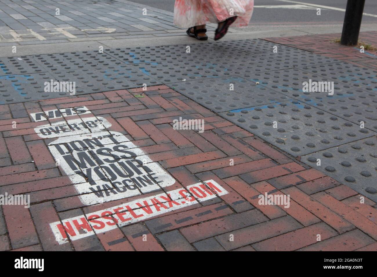 London, UK, 28 July 2021: In Windrush Square, Brixton, a painted sign on the pavement at a pedestrian crossing reminds anyone over the age of 18 that they are eligible for a coronavirus vaccination. Vaccine take-up is lower in big cities than in the rest of the country. Anna Watson/Alamy Live News Stock Photo
