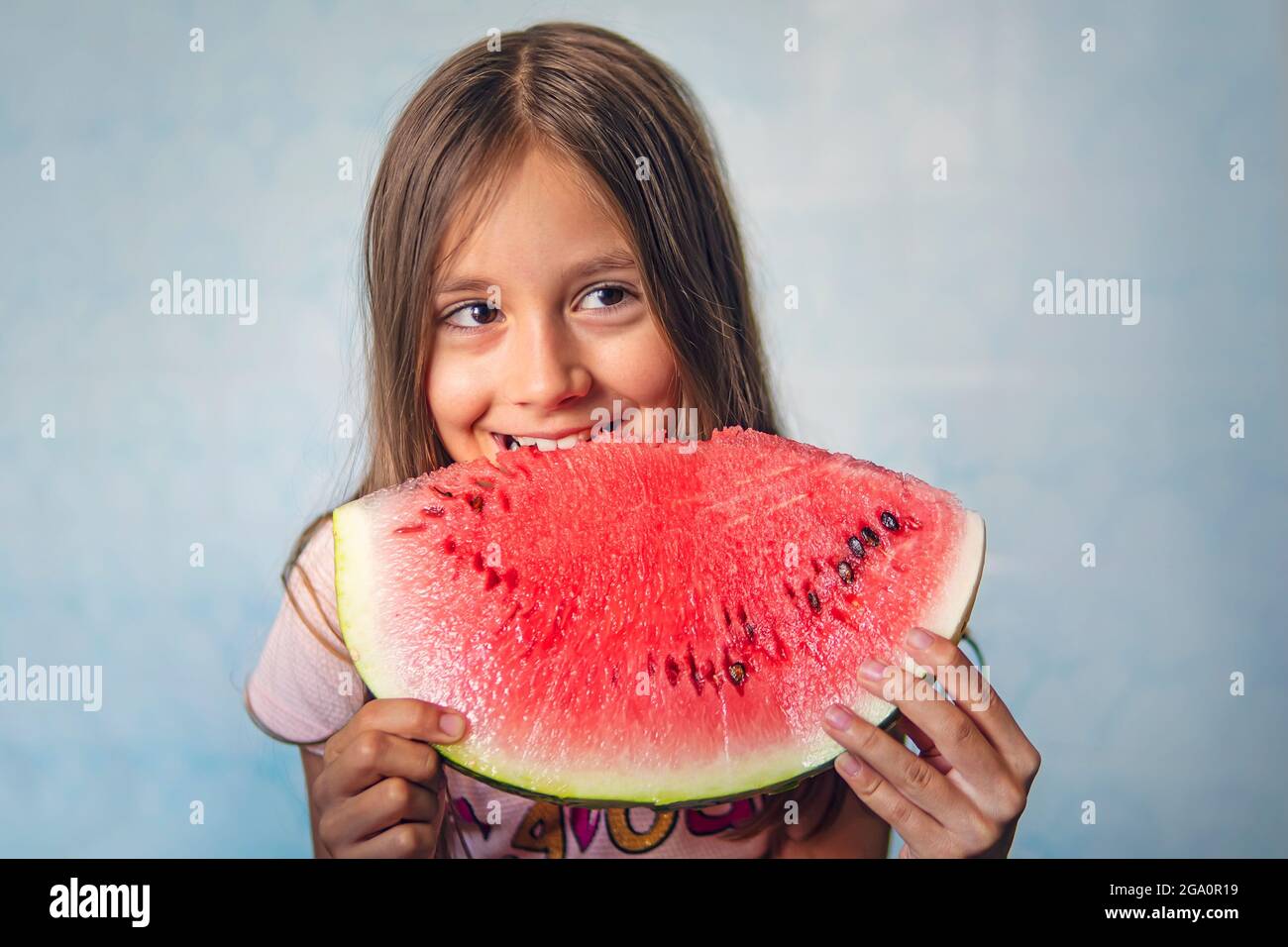 Happy toddler eating melon. A little girl child bites a large piece of watermelon on a blue background. Stock Photo