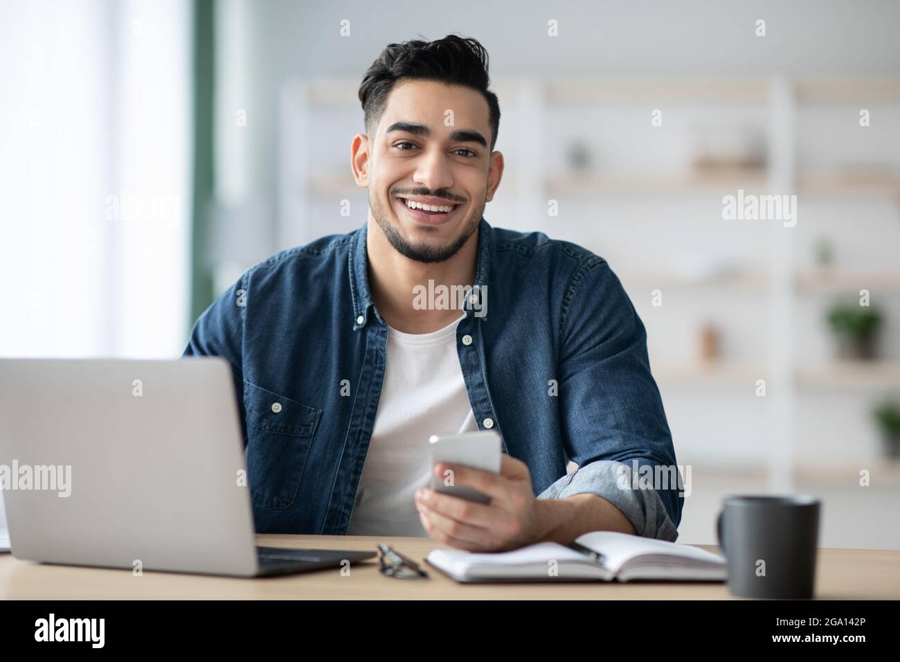 Smiling arab guy using smartphone while sitting at workdesk Stock Photo