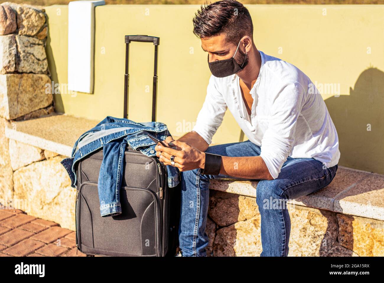 Solo traveller waiting for public transport sitting on bench wearing safe protective black face mask using smartphone to pass tedious waiting. Stylish Stock Photo