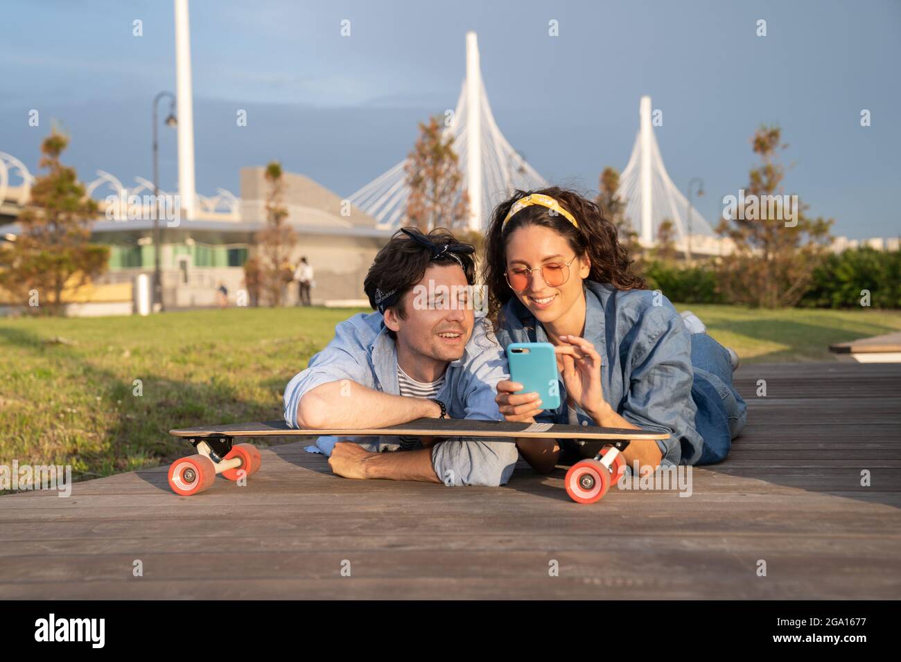 Young stylish girl show guy text message on smartphone lying on longboard in urban park outdoors Stock Photo