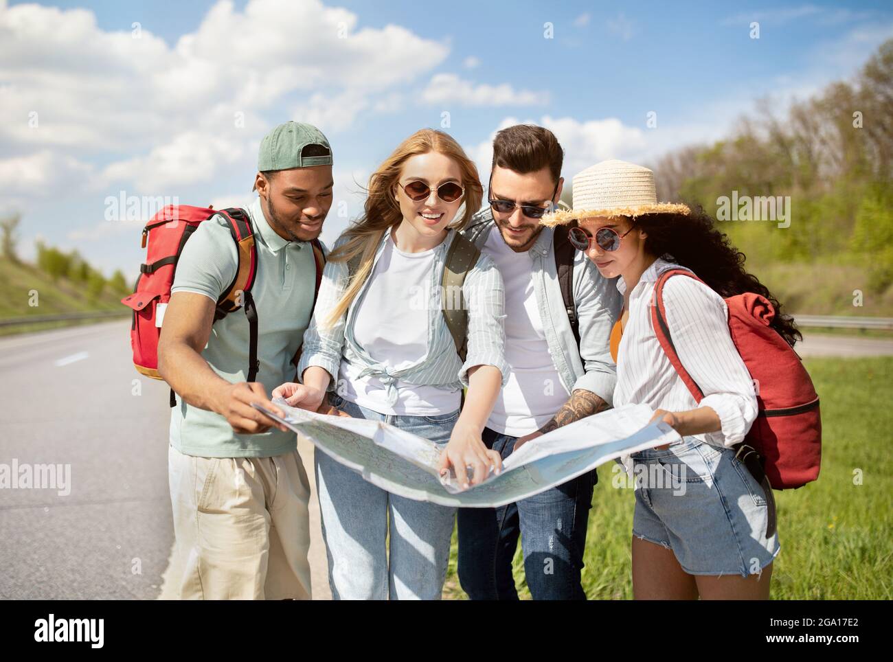 Group of best friends with backpacks checking map during road trip, hitchhiking on highway Stock Photo
