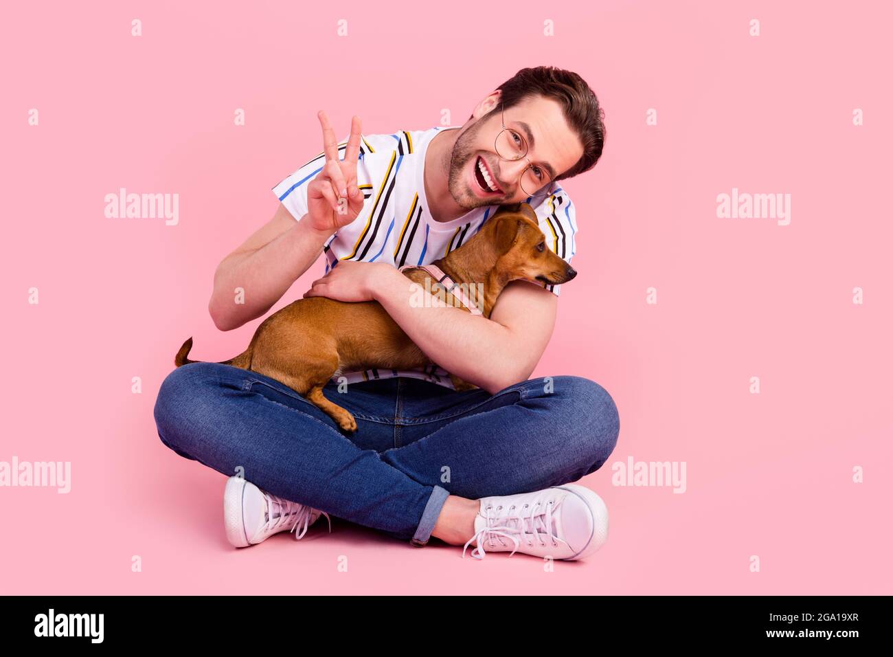 Full size photo of cheerful young positive man hug dig make v-sign sit ...