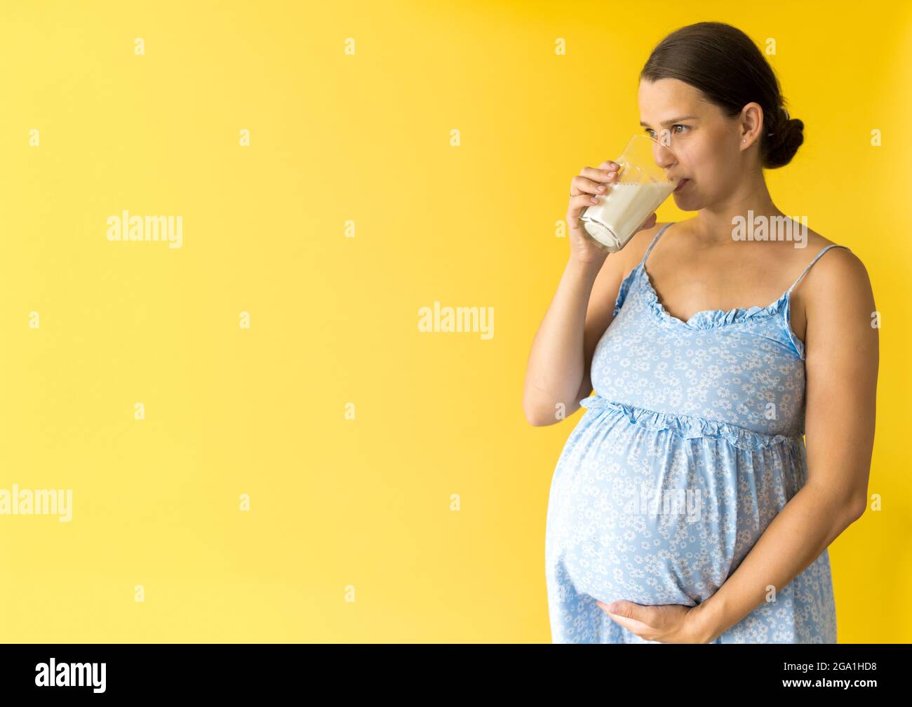 portrait cute beautiful pregnant young pretty woman in floral blue dress holds drink glass of milk rubs tummy on yellow background. Motherhood Stock Photo