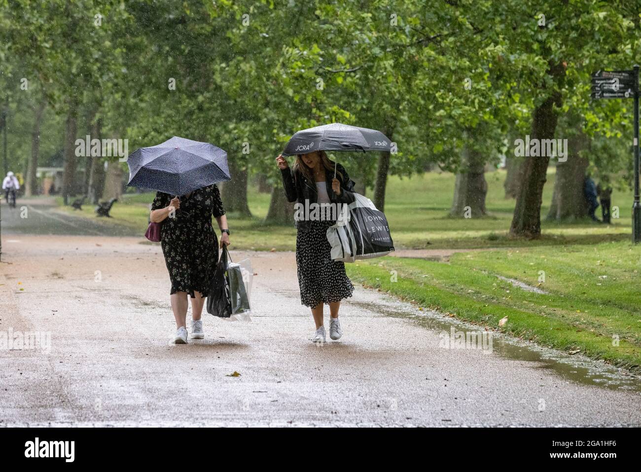 London, UK. July 28 2021: London, UK. July 28 2021: Heavy Rain Showers, Central London, England, UK Picture shows a couple taking cover underneath their umbrellas fas they make their way through the the rain in Hyde Park after some shopping as heavy rain showers sweep across Central London and southern parts of England. Credit: Clickpics/Alamy Live News Stock Photo