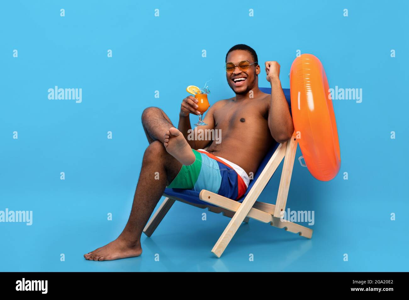 Full length of joyful black guy with tropical cocktail chilling in lounge chair, making YES gesture over blue background Stock Photo
