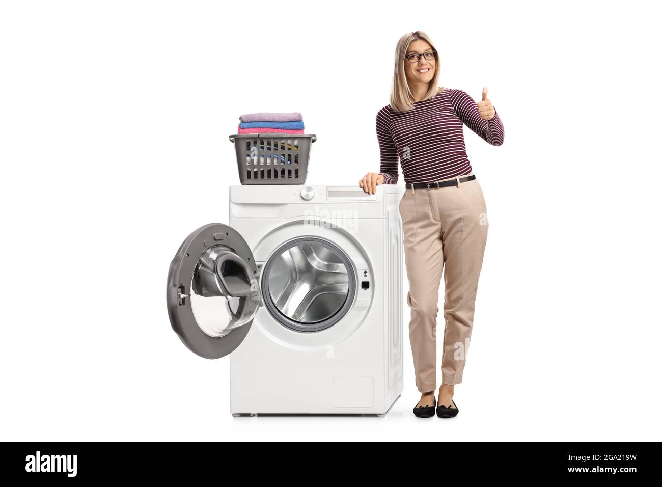 Full length portrait of a blond woman leaning on a washing machine with folded clothes and showing thumbs up isolated on white background Stock Photo