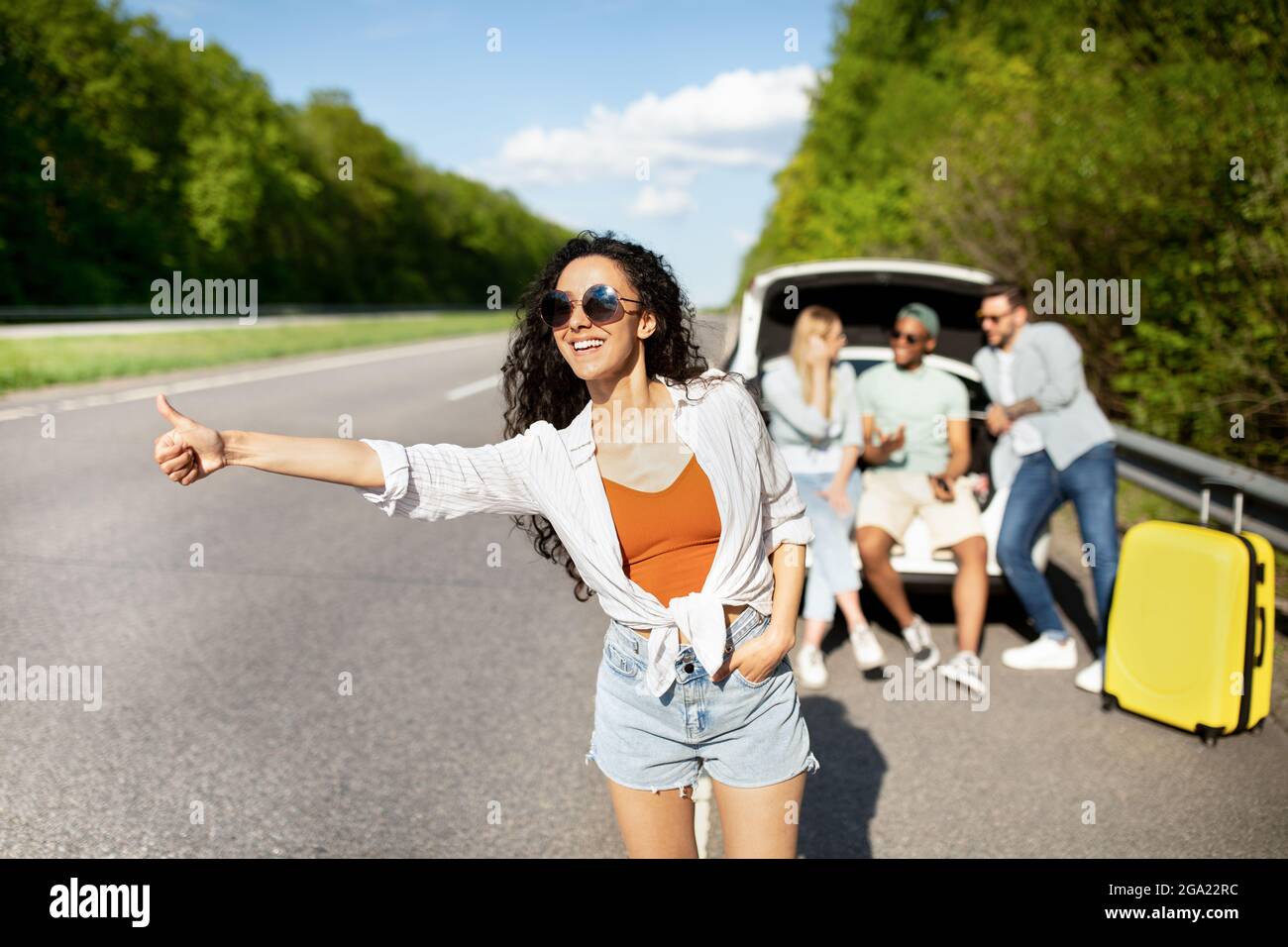 Young lady traveling with her friends, stopping car, hitchhiking on highway, needing help with broken vehicle Stock Photo
