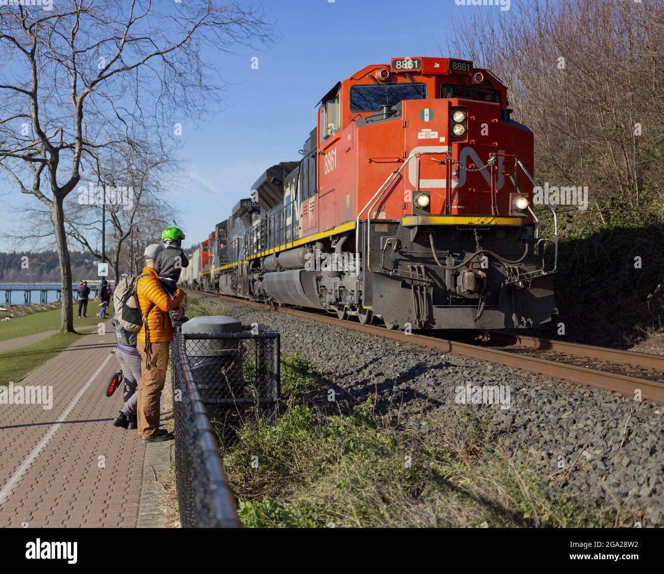 A young family stands on a walking trail on the BC coast watching a freight train pass on the tracks; White Rock, British Columbia, Canada Stock Photo