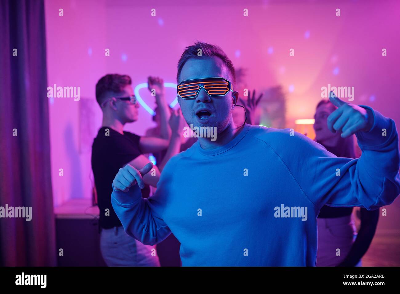 Young serious man in casualwear and disco eyeglasses looking at you while standing in front of his friends dancing together at home party Stock Photo