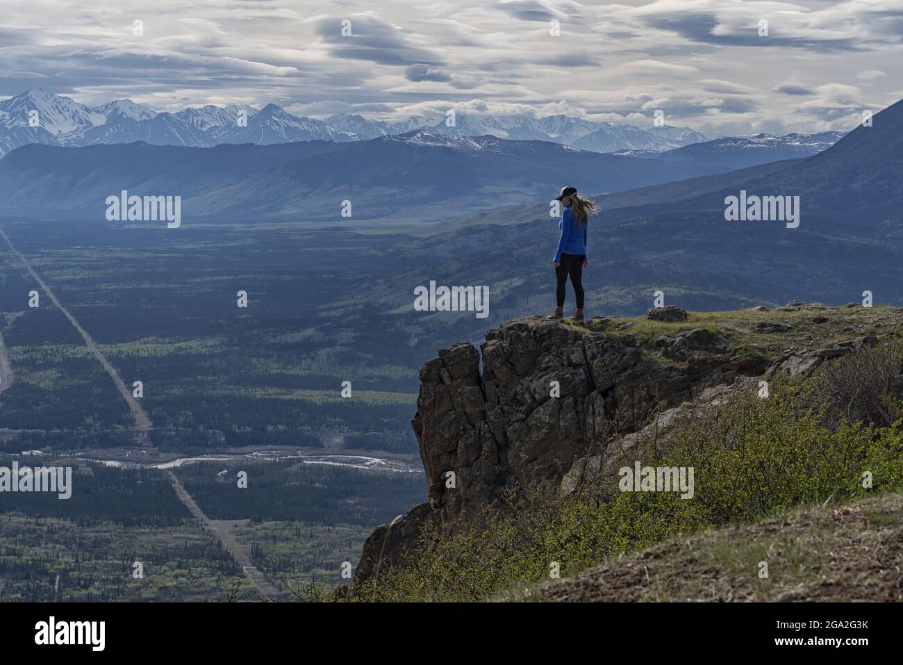 Woman standing on the edge of a mountain cliff looking out at the vista of the majestic mountain ranges and valley below with a cloudy sky on a sun... Stock Photo