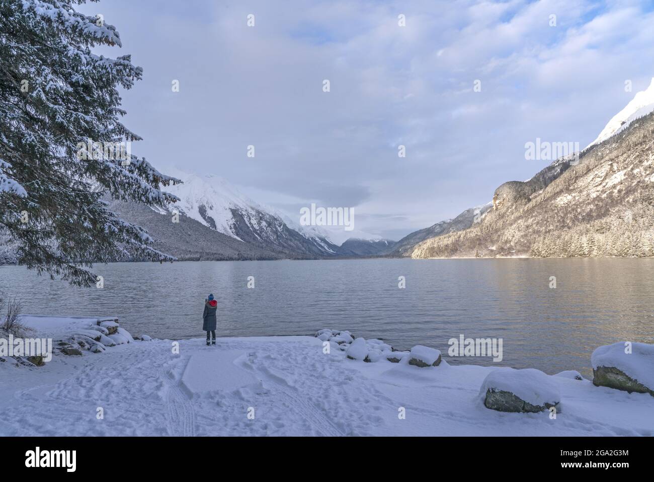 View from behind of a woman standing along the shore of a lake looking at the snow covered mountains; Haines, Alaska, United States of America Stock Photo