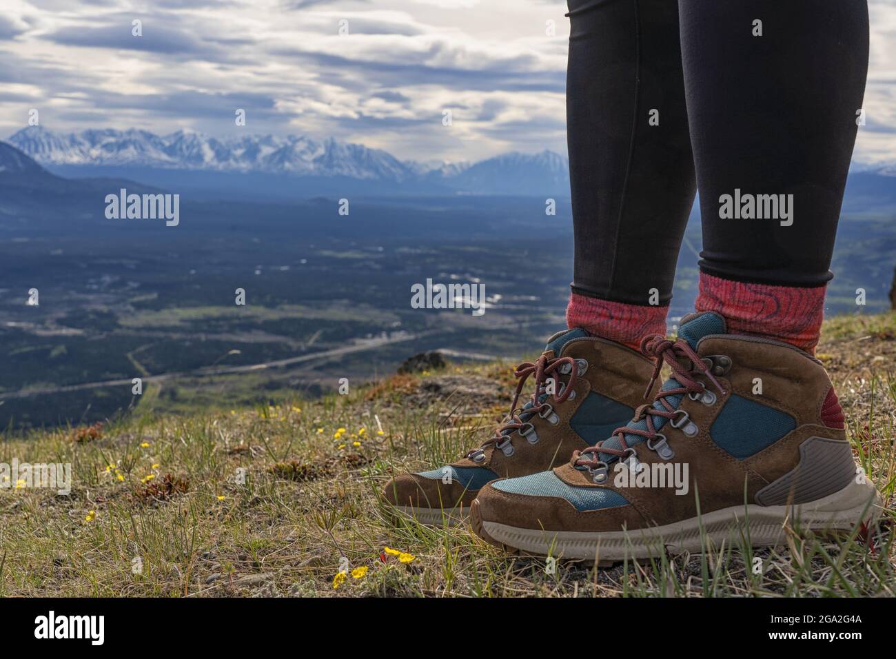 Close-up of woman's feet wearing hiking boots, standing on a mountaintop with a vista of the mountain ranges and valley below in the distance on a ... Stock Photo