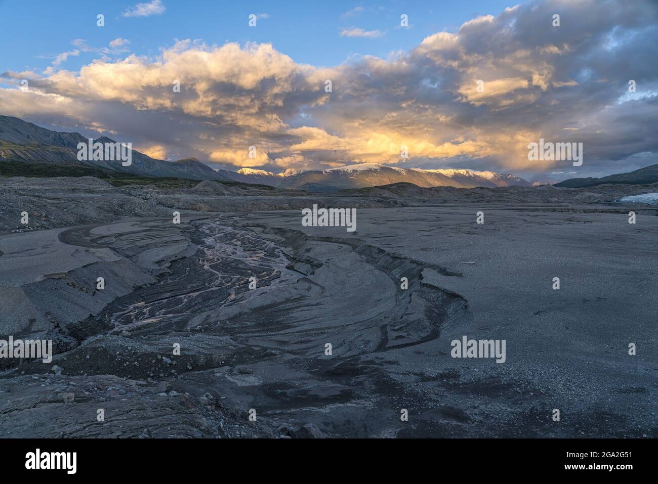 The setting sun lights up the clouds over the distant peaks of Kluane National Park, Yukon. A small stream flows in the foreground. Situated in the... Stock Photo