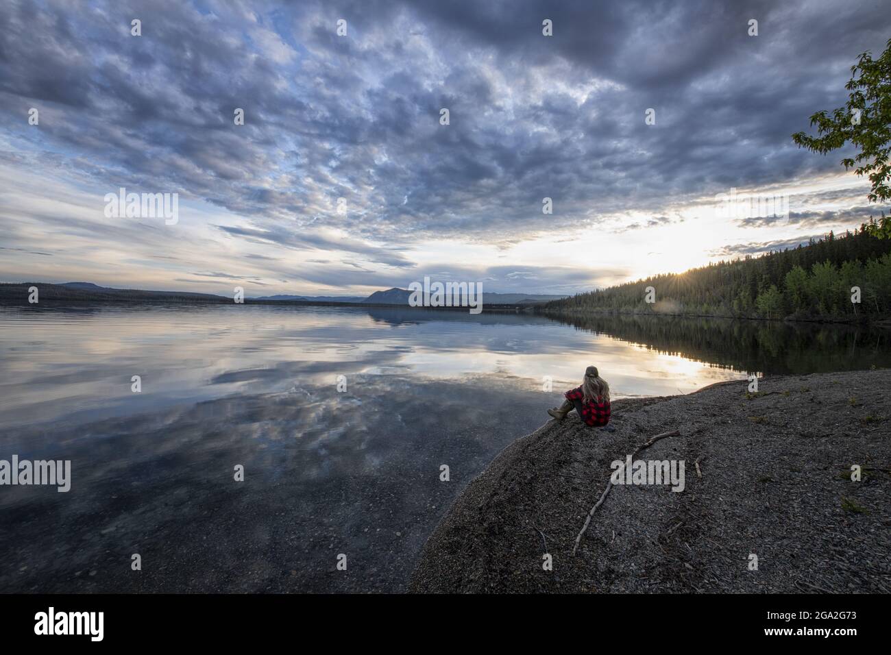 View taken from behind of a woman sitting alongside Little Salmon Lake at the water's edge enjoying the view during the late afternoon in the tradi... Stock Photo