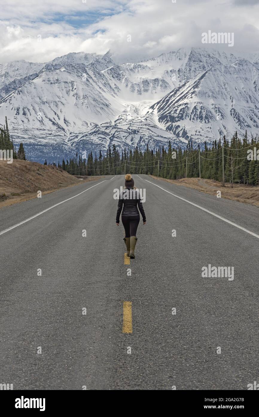 View taken from behind of a woman traveler walking towards the mountains in the middle of the Alaska Highway, situated in the Traditional Territory... Stock Photo