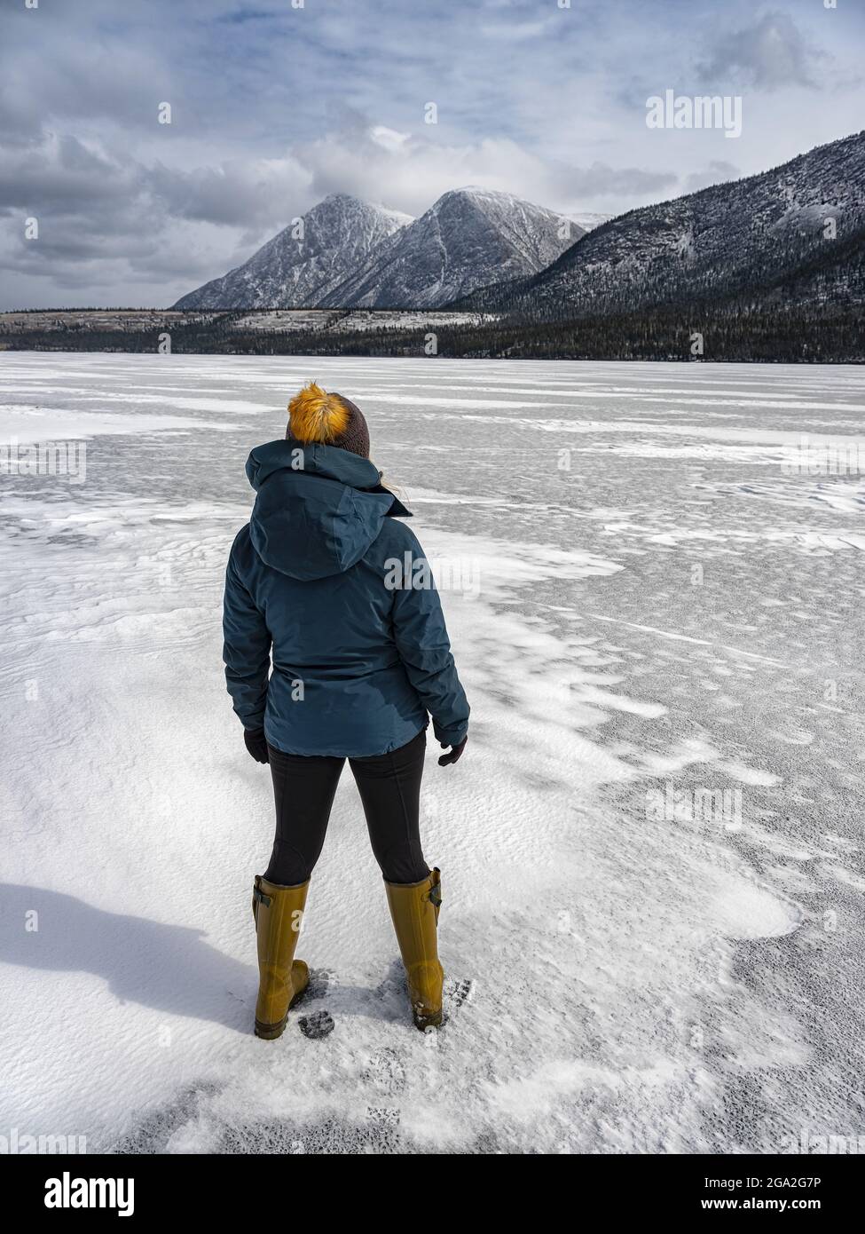 View taken from behind of a woman traveler standing on the frozen Kusawa Lake near Whitehorse; Yukon, Canada Stock Photo