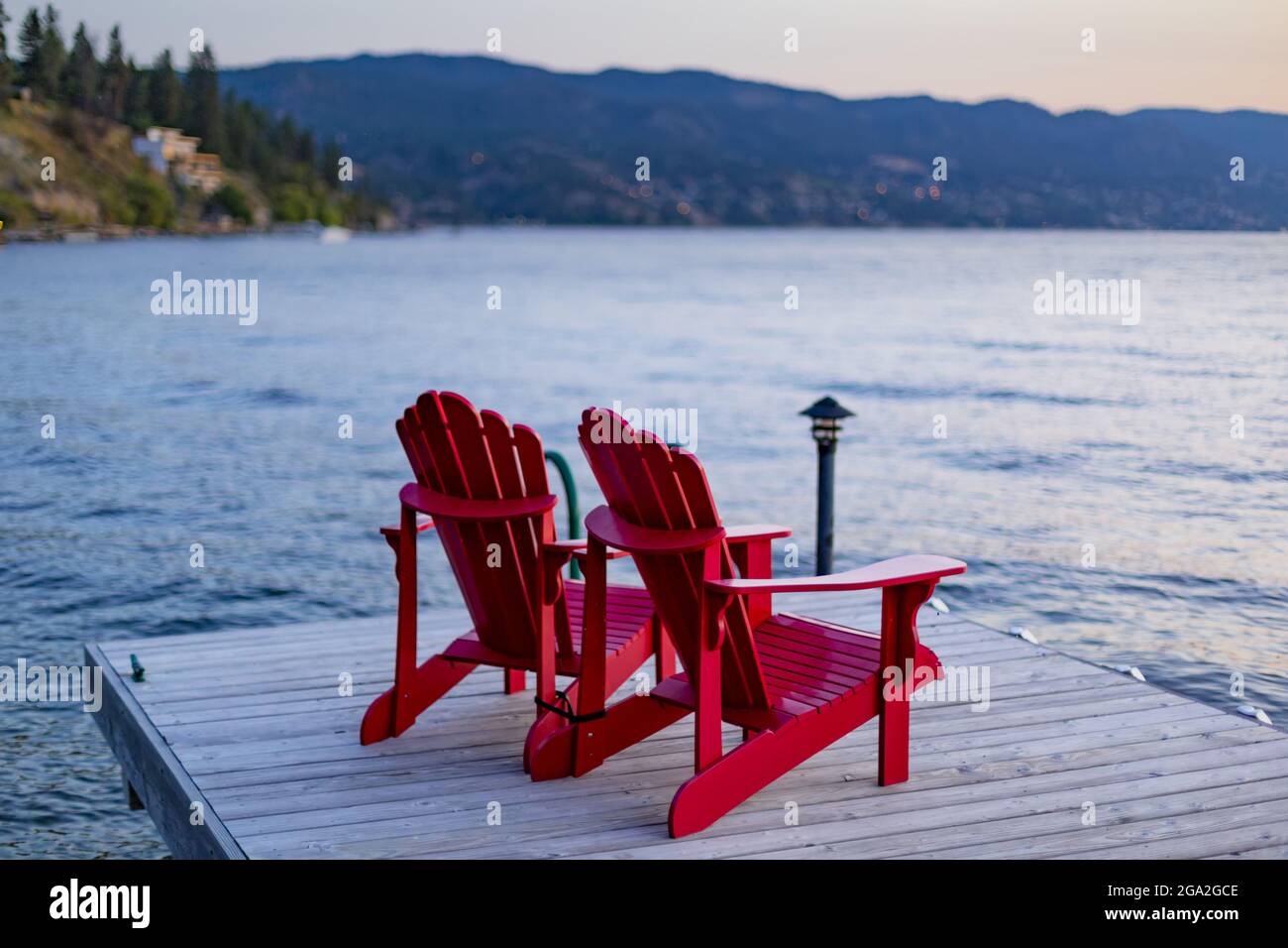 Two Adirondack chairs sit on a wooden dock on Okanagan Lake at sunset; British Columbia, Canada Stock Photo