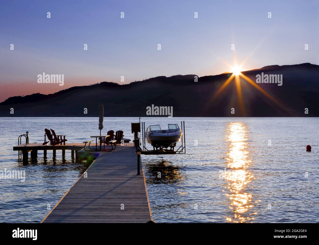 Adirondack chairs and a picnic table sit on a wooden dock with a boat and lift on Okanagan Lake at sunset; British Columbia, Canada Stock Photo