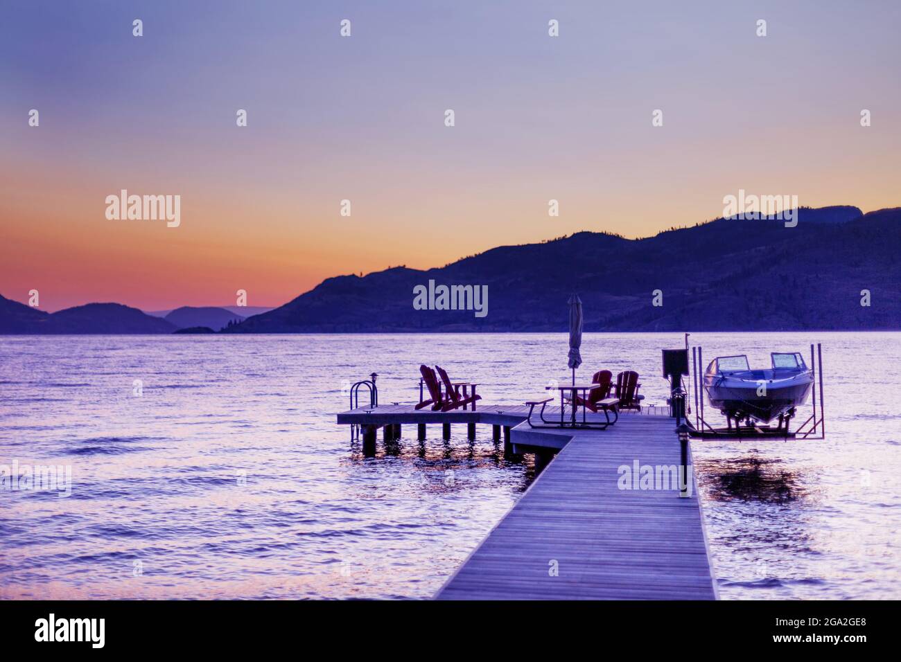 Adirondack chairs and a picnic table sit on a wooden dock with a boat and lift on Okanagan Lake at sunset; British Columbia, Canada Stock Photo