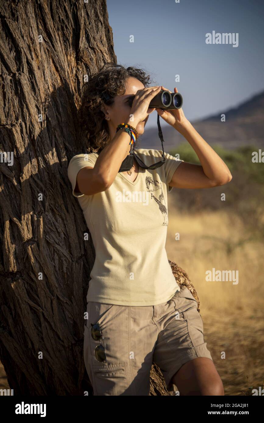 Portrait of a woman leaning on a tree trunk wearing shorts and looking through binoculars into the bright sunlight into the distance at the Gabus G... Stock Photo