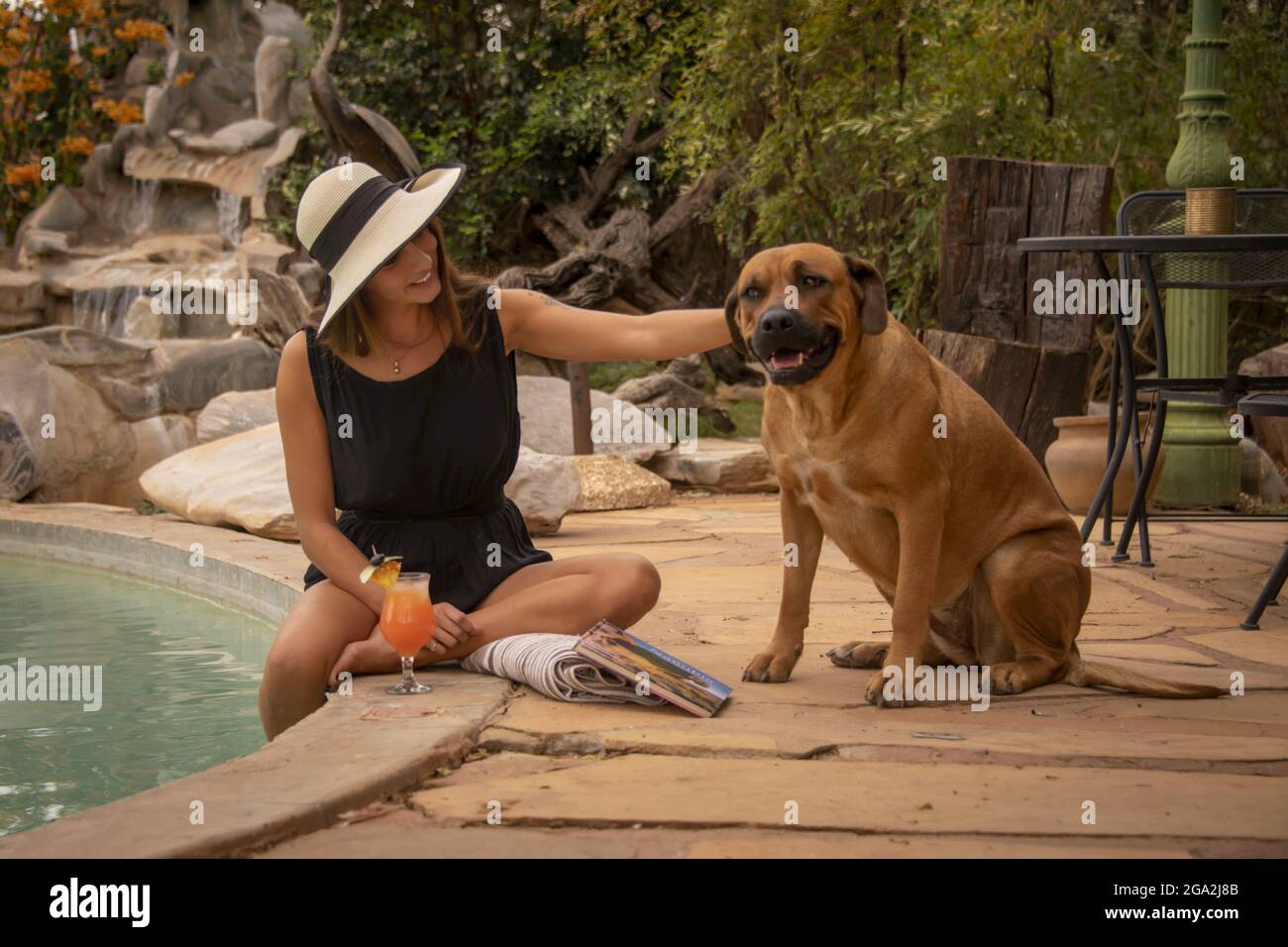 Woman wearing sunhat sitting by a swimming pool and petting a dog (Canis lupus familiaris) at the Gabus Game Ranch; Otavi, Otjozondjupa, Namibia Stock Photo