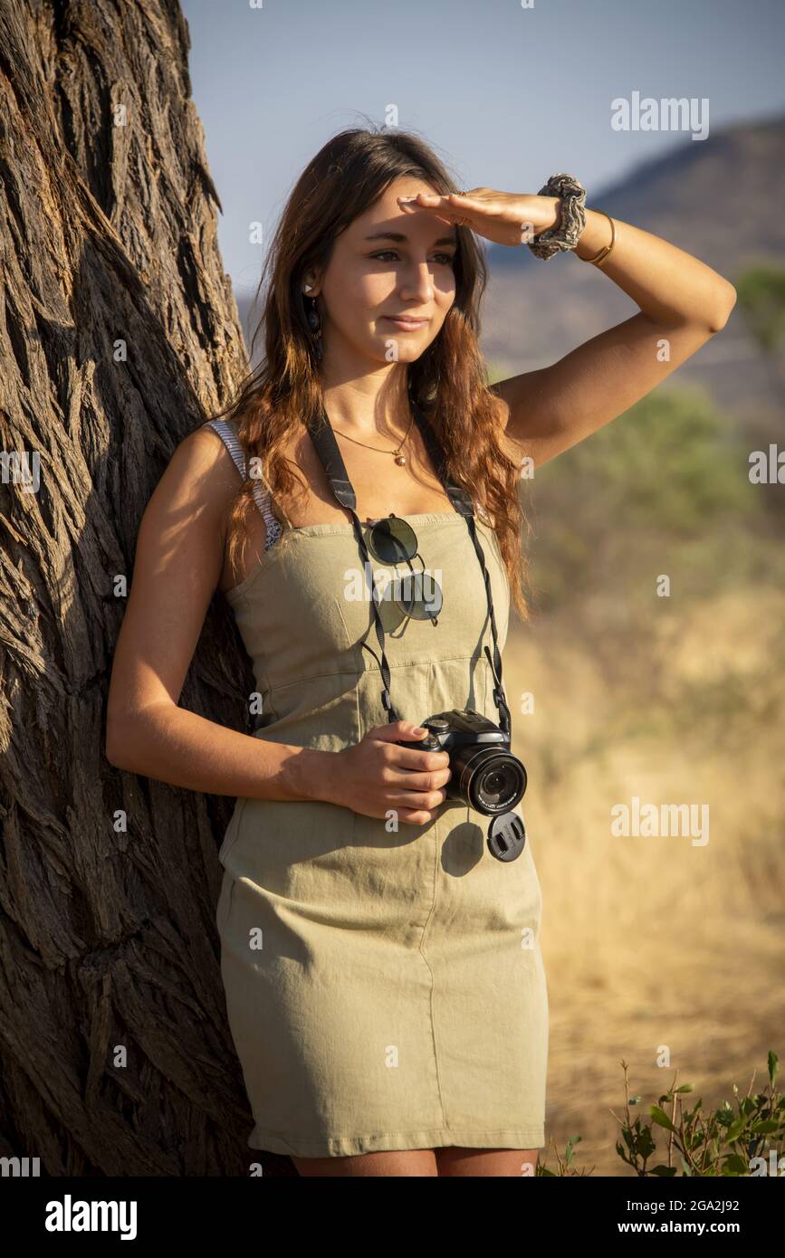 Portrait of a woman leaning on a tree trunk wearing a sundress and holding a camera while shading her eyes against the bright sunlight and looking ... Stock Photo