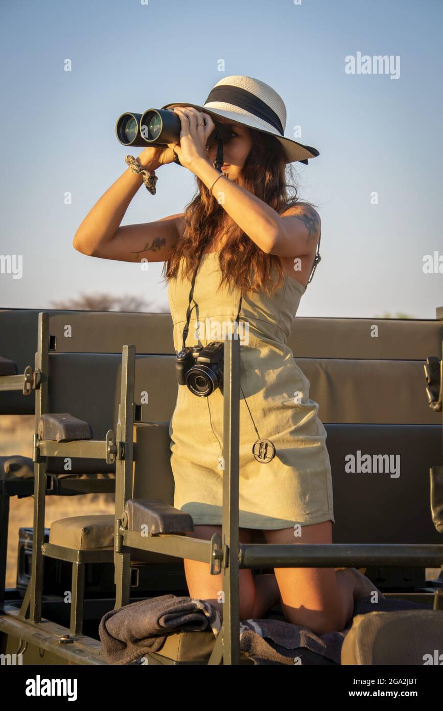 Close-up of a woman kneeling in a jeep on safari, wearing a straw hat with a camera hanging around her neck and using binoculars to look out onto t... Stock Photo
