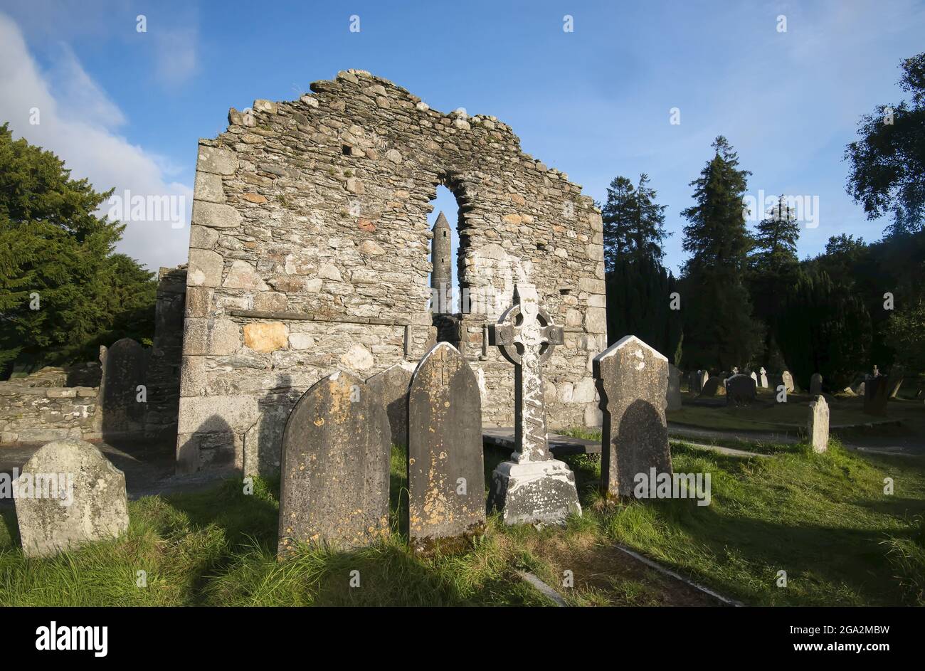 Tombstones and Celtic Cross memorial with the Round Tower seen through the lancet window of the ruins at Glendalough (or The valley of the Two Lake... Stock Photo