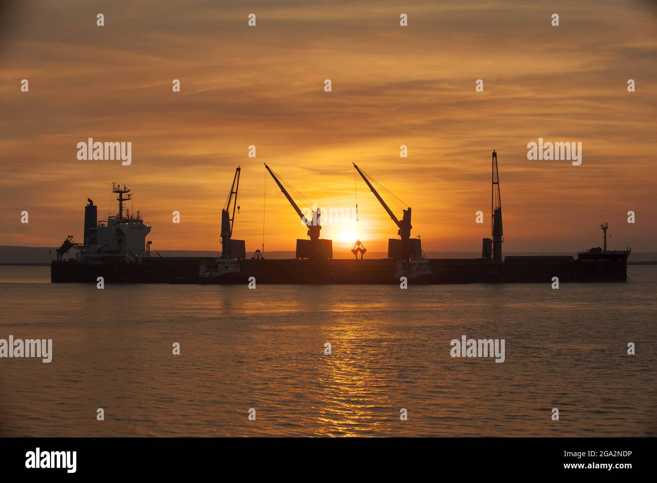 The sun sets behind a cargo vessel as it loads iron ore off the coast of Western Australia; Western Australia Stock Photo