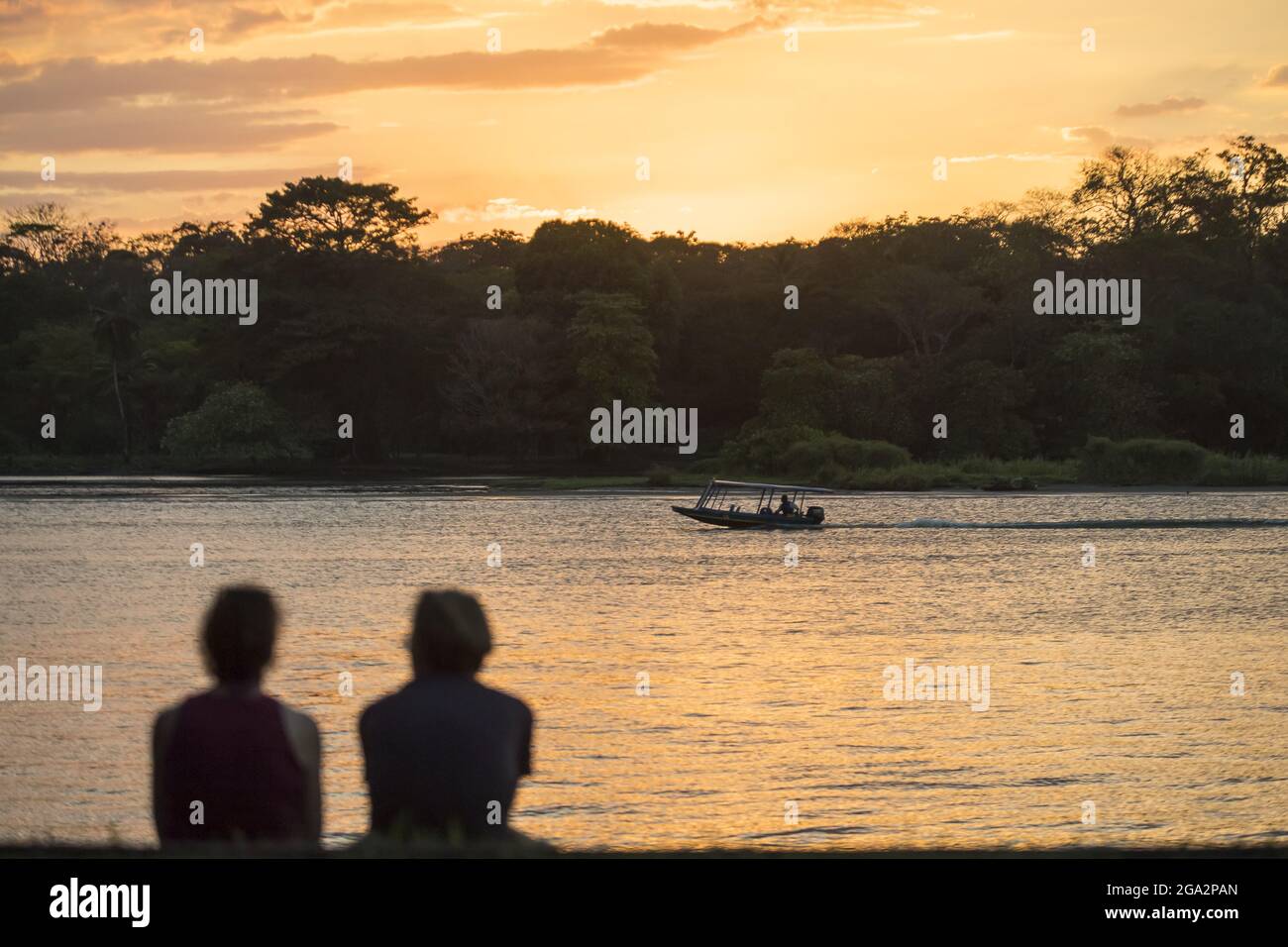 View from behind of a silhouette of tourists sitting on the shore watching a golden sunset with a boat traveling the waterways that surround Tortug... Stock Photo