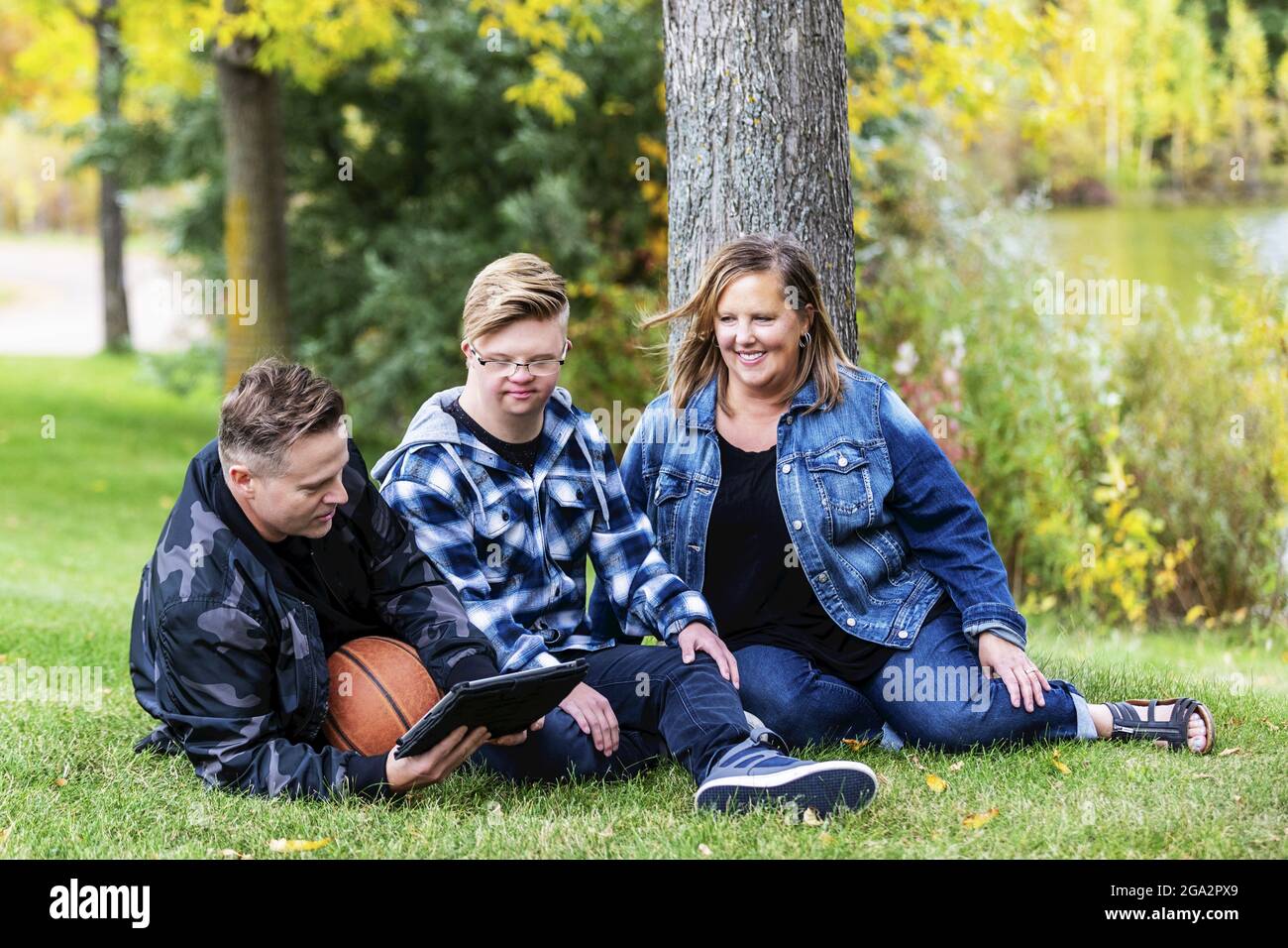 A young man with Down Syndrome sitting with his family and playing games on a tablet, while enjoying each other's company in a city park on a warm ... Stock Photo