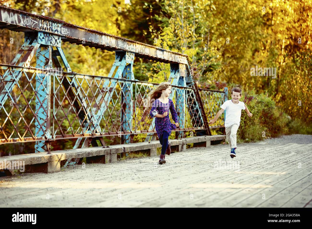 A brother and sister running across a bridge and spending quality time together outdoors in a city park during the fall season Stock Photo