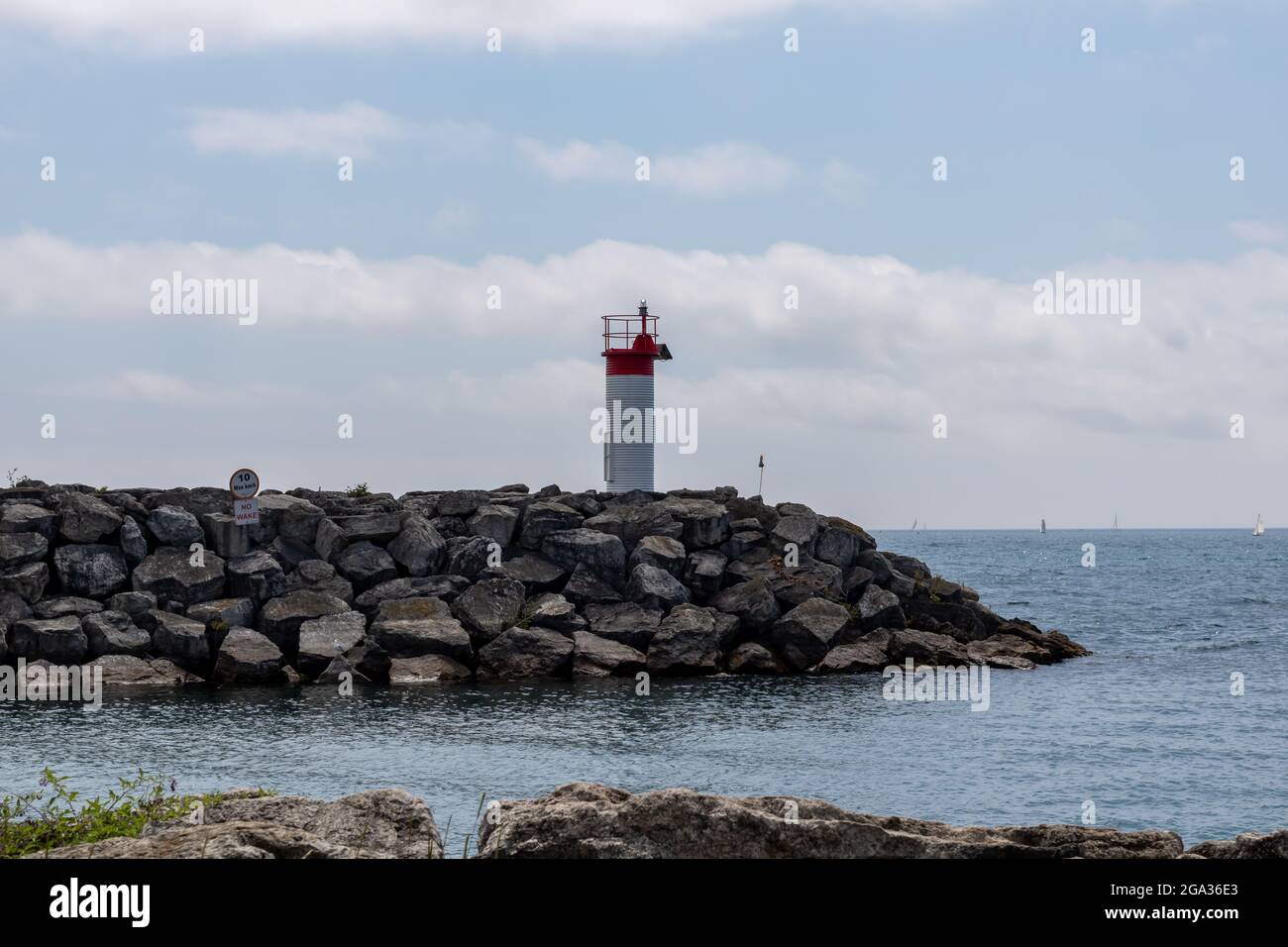 A small red and white lighthouse on a rocky pier at Lakefront Promenade Park, Mississauga, Lake Ontario. Sailboats in background. Stock Photo