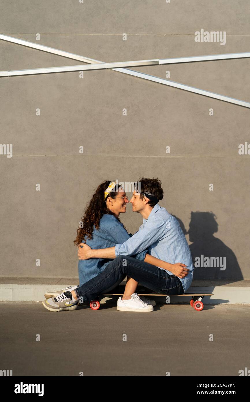 Trendy young couple sit on longboard on sunny city street happy smiling embrace enjoy time together Stock Photo