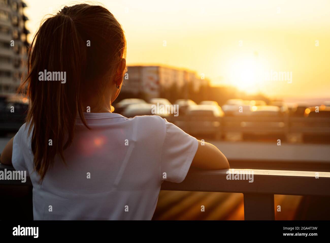Schoolgirl at back sunset. Teenage girl blonde with long hair in ponytail admires setting sun light and blurry city buildings close backside view Stock Photo