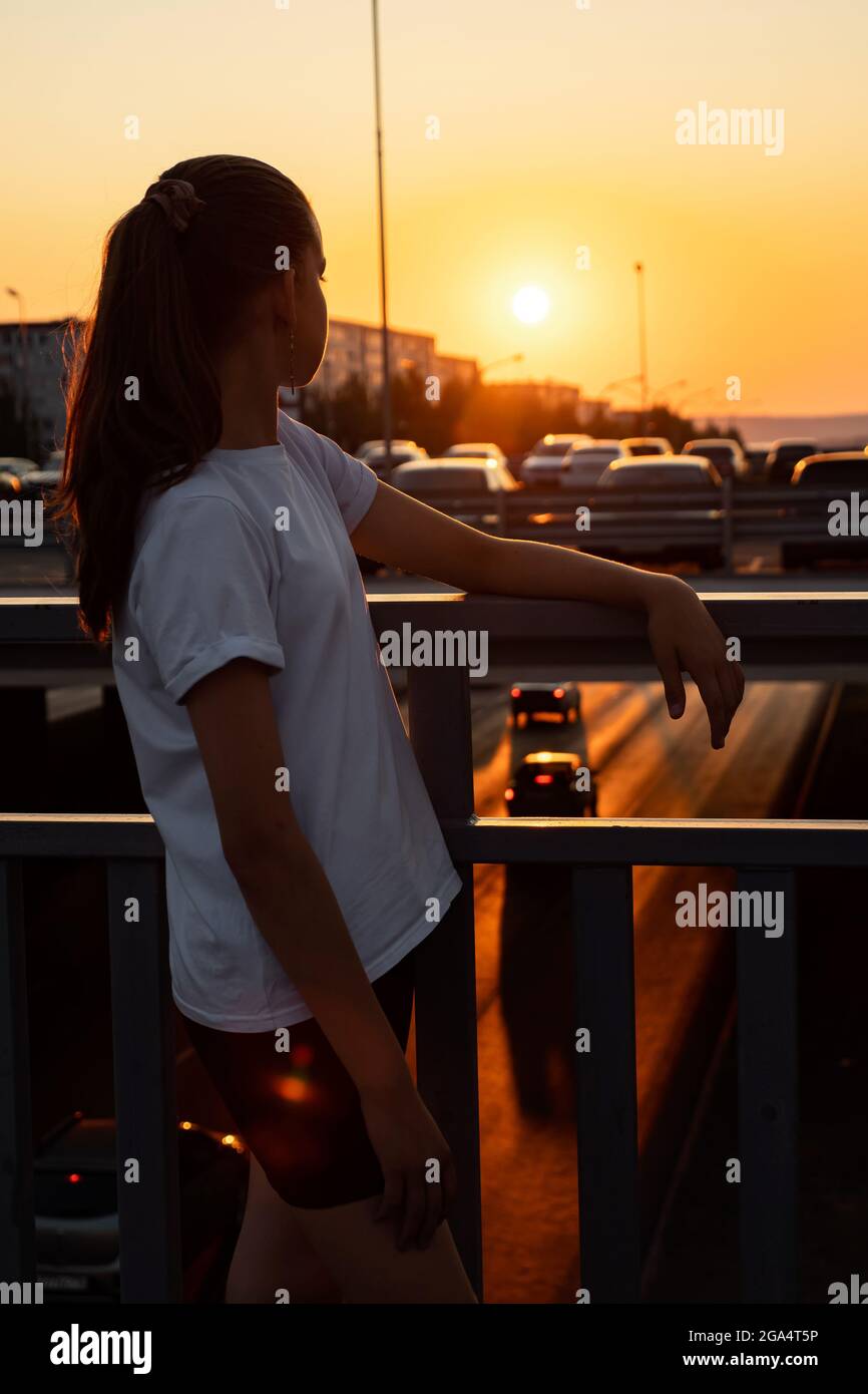 Teenage girl with long hair in ponytail in t-shirt looks at sunset, standing on overpass bridge backside view Stock Photo