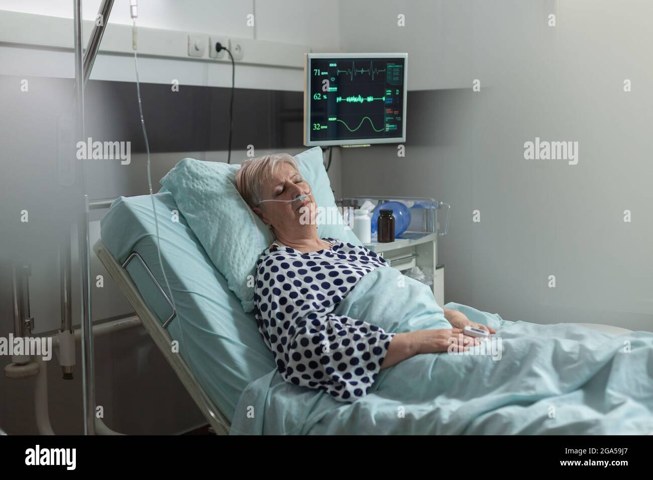 Senior woman patient in hospital room getting medicine through intravenous line from iv drip bag and breathing with help from oxygen mask laying in bed. Oxymeter attached on finger monitoring bpm. Stock Photo